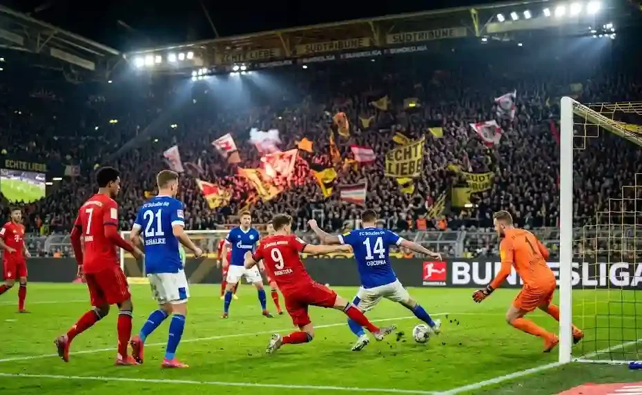 Players in red and blue jerseys competing for the ball near the goal in a packed Bundesliga stadium under floodlights.