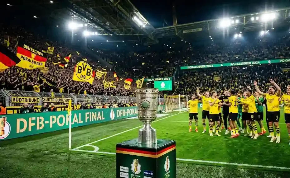 A detailed silver DFB-Pokal trophy resting on a podium near the pitch with football players celebrating victory in front of a cheering crowd in a illuminated German stadium.