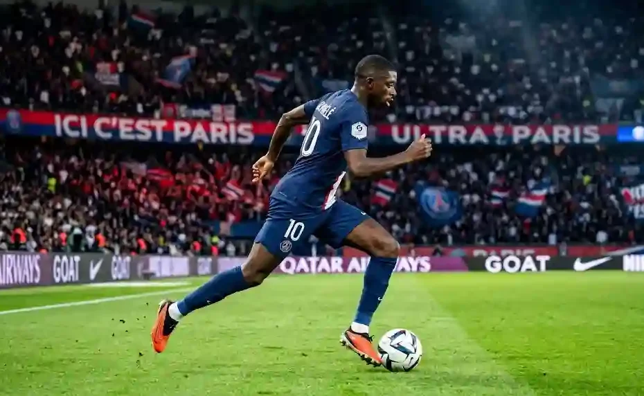 Ousmane Dembélé in navy-blue PSG jersey sprinting with the soccer ball at Parc des Princes stadium under bright lights.